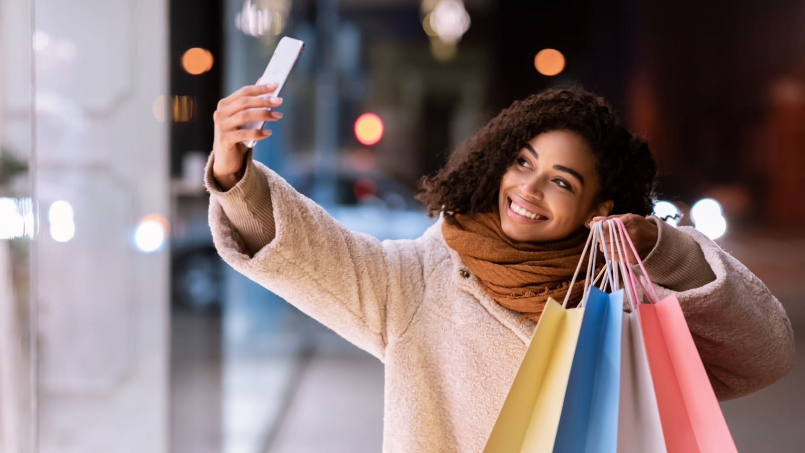 It's Shopping Time. Happy african american woman taking selfie standing with colorful bags near center, walking in the evening outdoors, posing at camera of her mobile phone. Entertainment Concept
