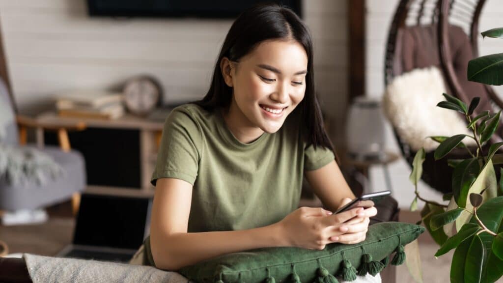 Smiling korean girl chatting on mobile phone, sitting at home in living room with smartphone and laughing happy