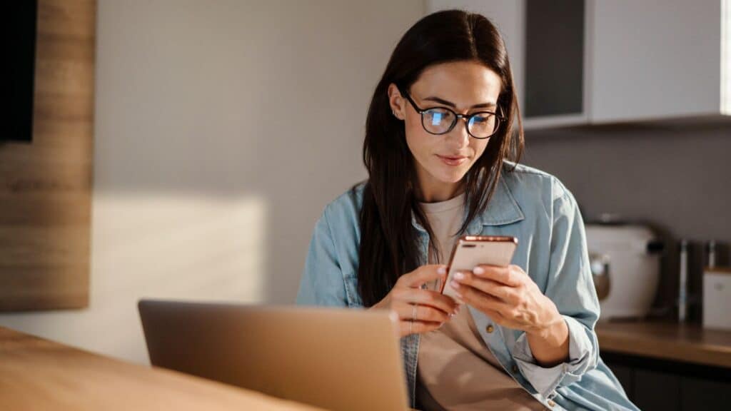 Woman using a smartphone while working with a laptop at home.