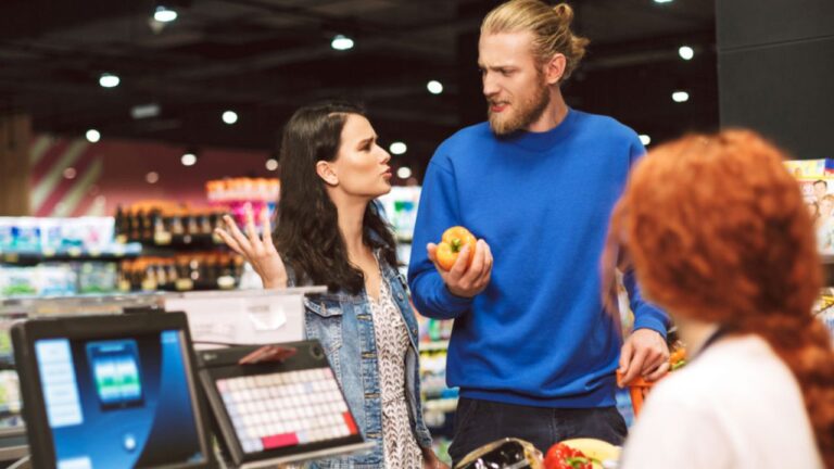 Young couple standing near cashier desk emotionally discussing something buying products together in modern supermarket