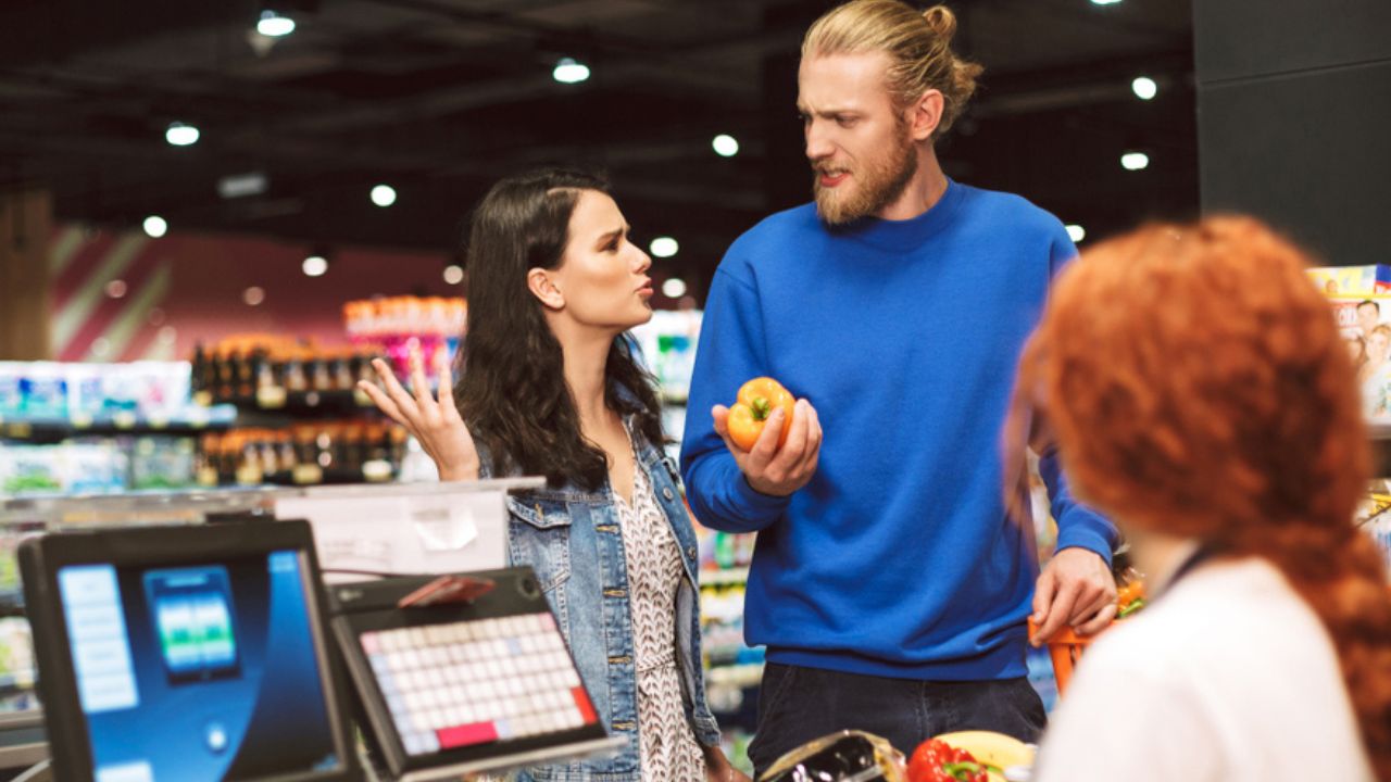 Young couple standing near cashier desk emotionally discussing something buying products together in modern supermarket