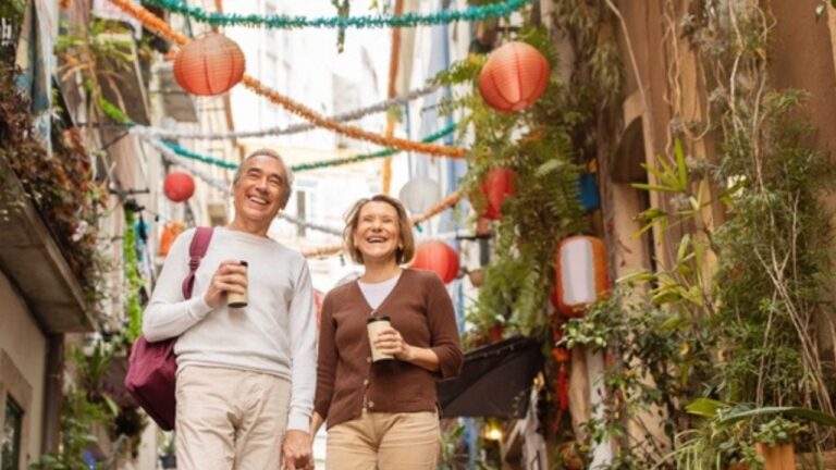 Cheerful Senior Travelers Couple Enjoying Walking Tour Holding Hands And Drinking Coffee On A Street In Lisbon Outside. Tourists Traveling In Europe After Retirement. Low Angle, Full Length Shot