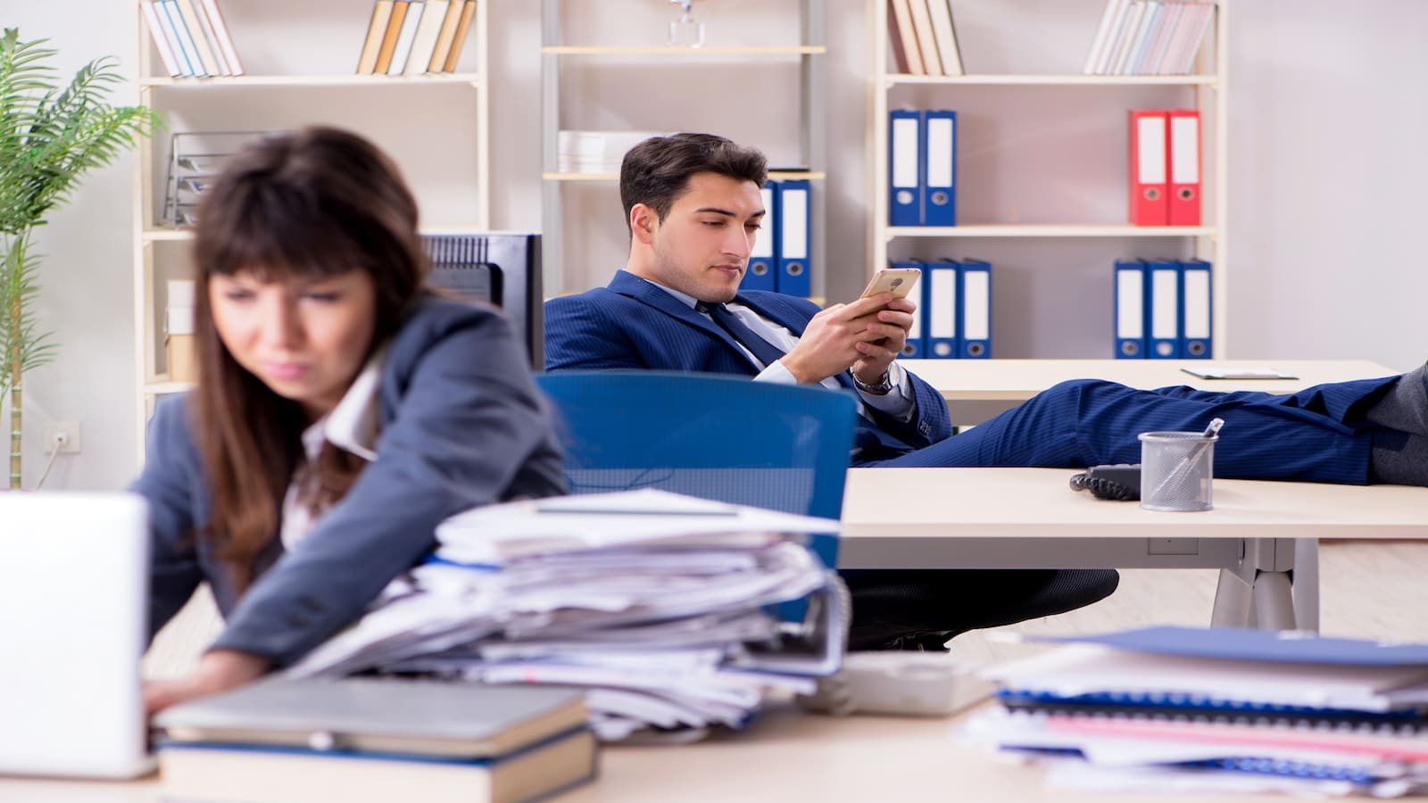 Man being selfish looking at phone and woman working hard in office.