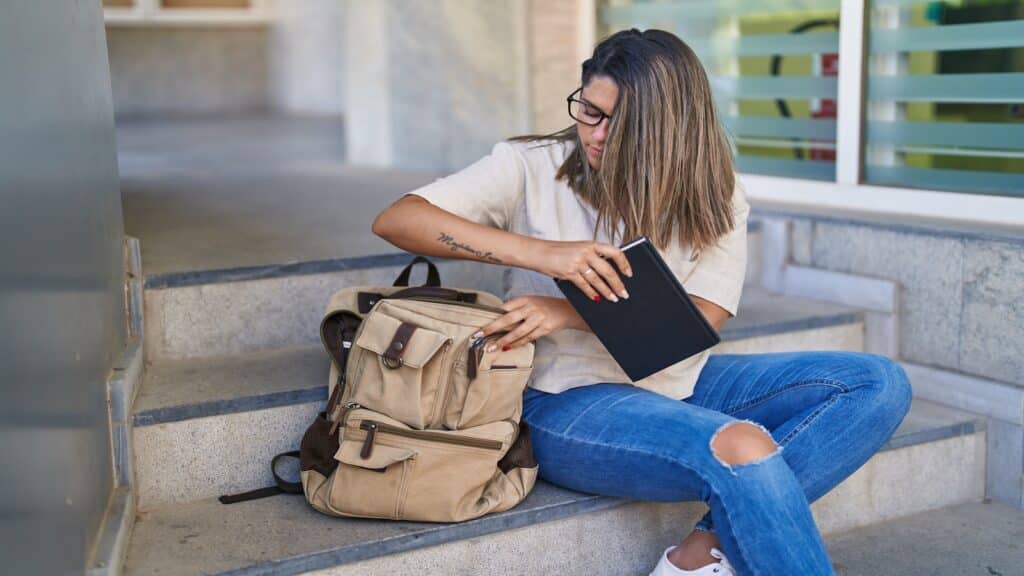 Young woman putting book notebook in bag sitting on stairs.