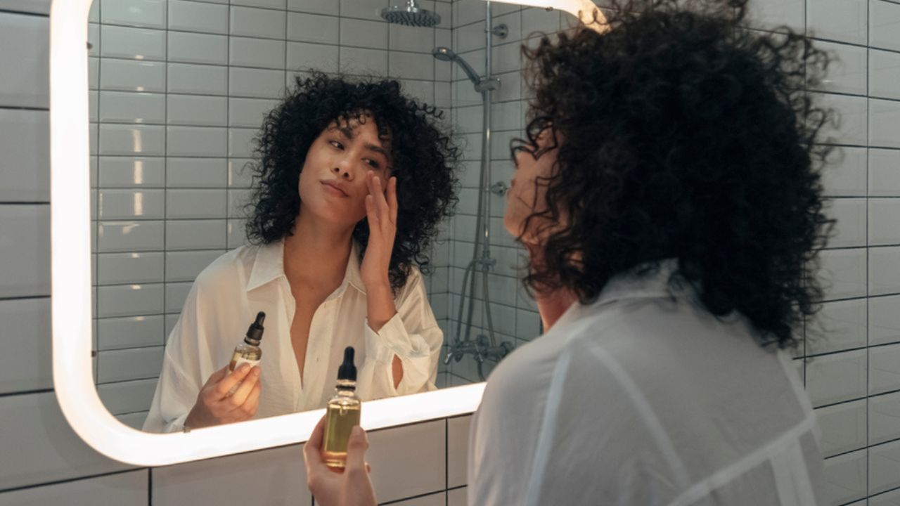 Young mixed race female gently applying face oil serum with fingers in modern bathroom. Women skincare and beauty concept.