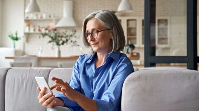 Relaxed mature old 60s woman, older middle aged female customer holding smartphone using mobile app, texting message, search ecommerce offers on cell phone technology device sitting on couch at home.