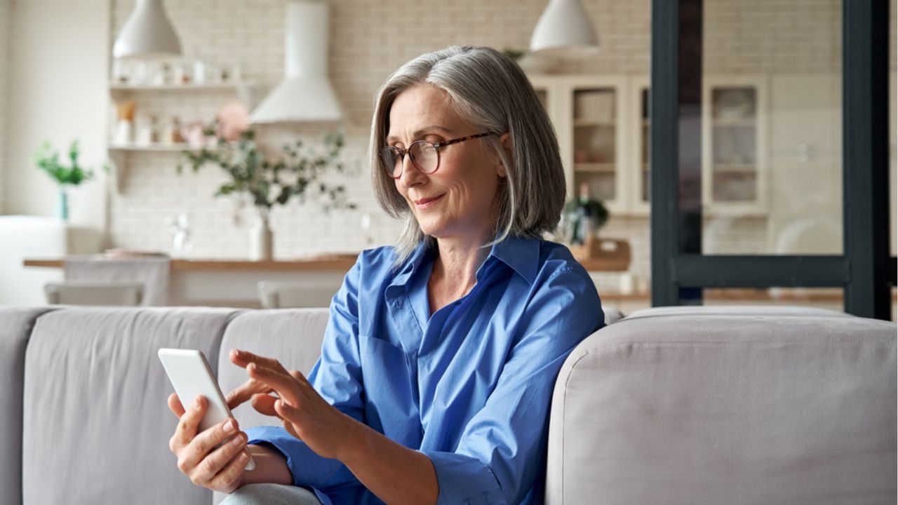 Relaxed mature old 60s woman, older middle aged female customer holding smartphone using mobile app, texting message, search ecommerce offers on cell phone technology device sitting on couch at home.