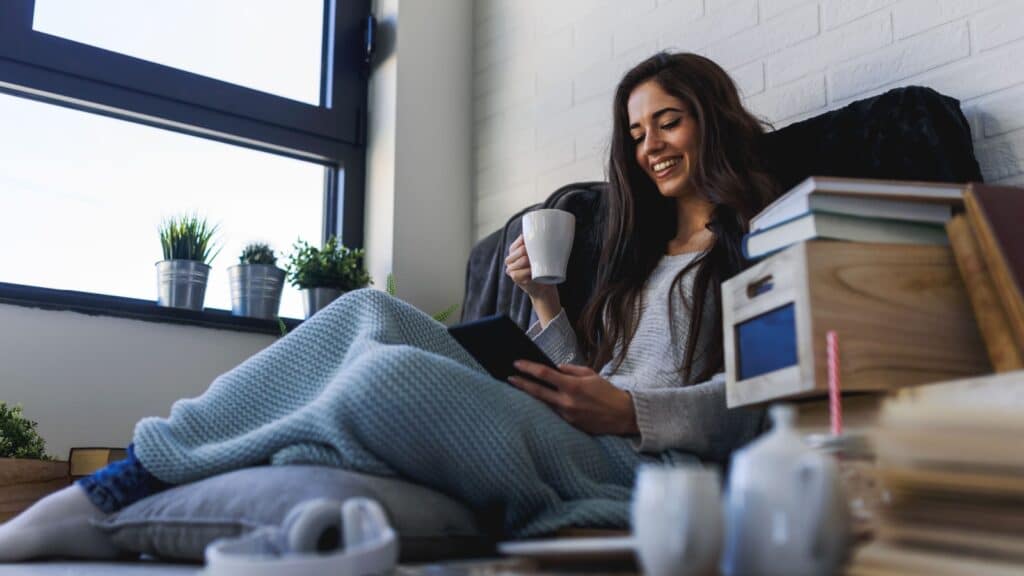 Beautiful young woman reading eBook reader at home surrounded by books.