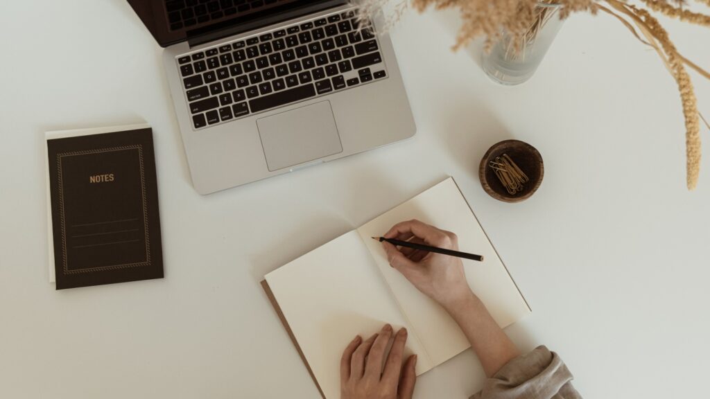 Flatlay of women's hands writing in a notebook sheet. Comfortable home office workspace interior design. Work at home. Laptop computer, notebook, pampas grass bouquet on table. Flat lay, top view.