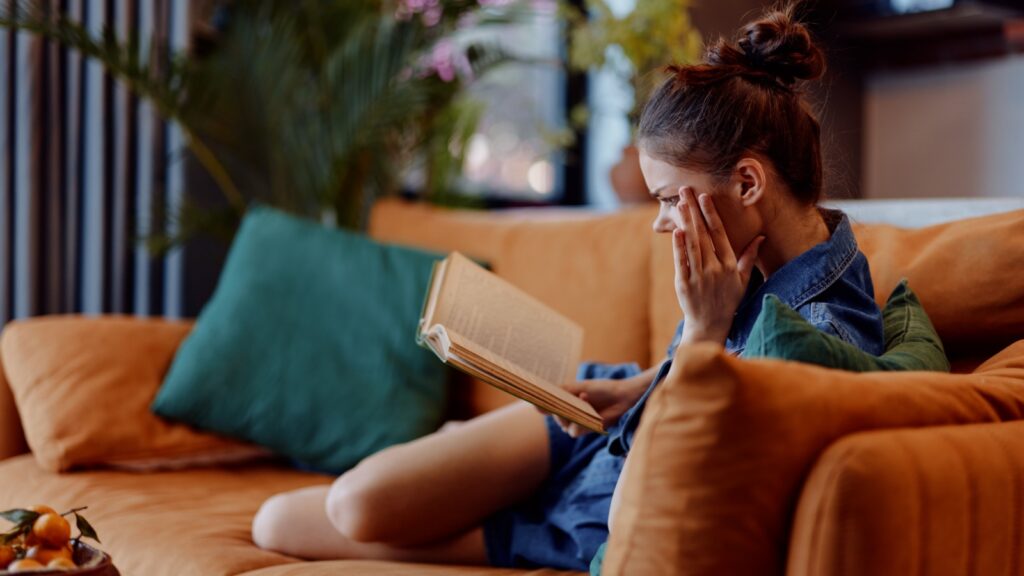 Relaxing at Home Woman Sitting on Couch with Eyes Closed and Book in Front of Her, Enjoying Peaceful Moment.