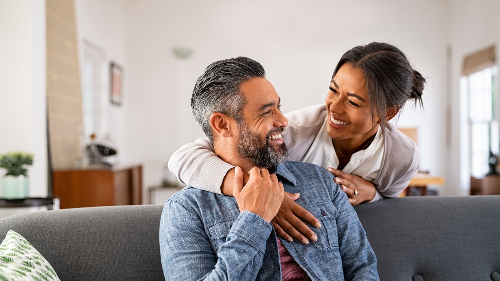 Smiling woman hugging her husband on the couch from behind in the living room.