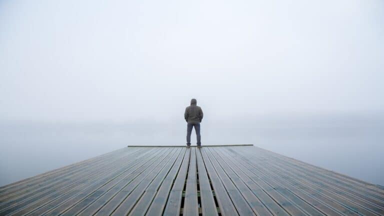 Young man standing alone on edge of footbridge and staring at lake with mist over water and foggy air.