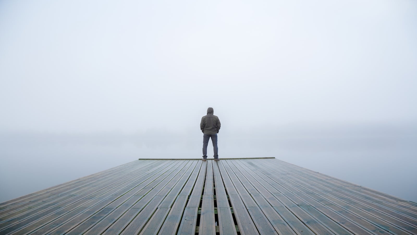 Young man standing alone on edge of footbridge and staring at lake with mist over water and foggy air.