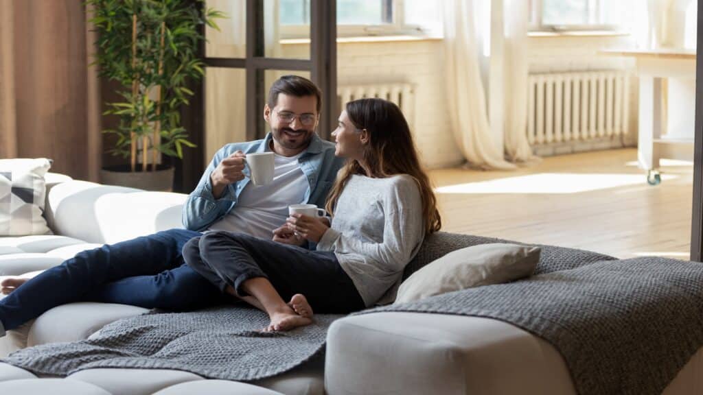 Loving couple relaxing on cozy sofa in living room, drinking hot beverages, tea or coffee, happy smiling man and woman holding white cups sitting on couch, chatting, talking, spending lazy weekend.