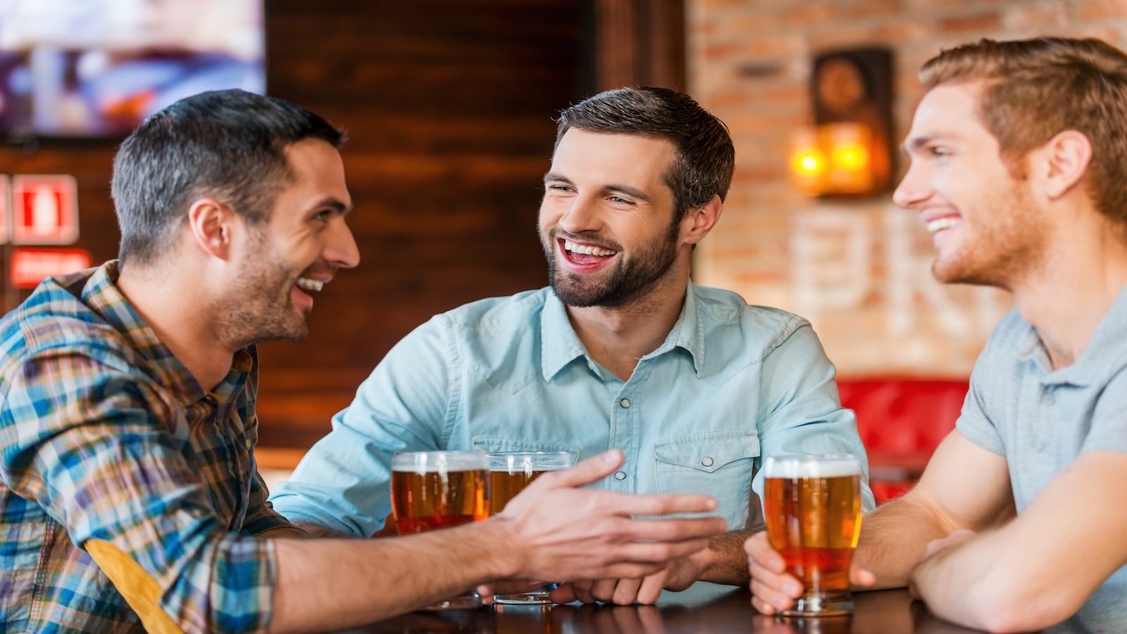 Three happy young men in casual wear talking and drinking beer while sitting in bar together.