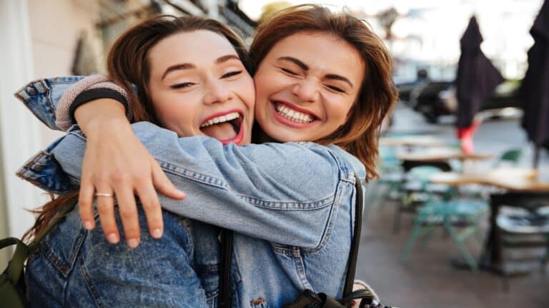 Close-up photo of laughing woman friends hugging each other on city street.