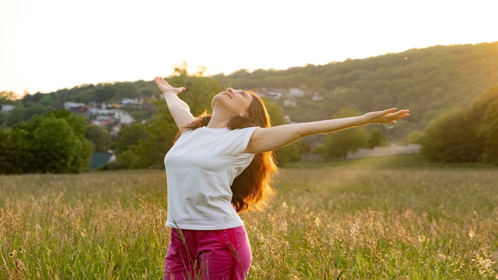 A woman stands in a field in nature with her hands raised.
