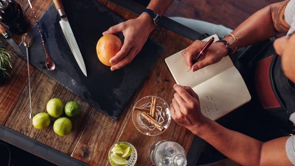 Barman accessories and ingredients on counter. Bartender holding a grapefruit on to the chopping board with another taking down notes. Men experimenting with creating cocktail drink.