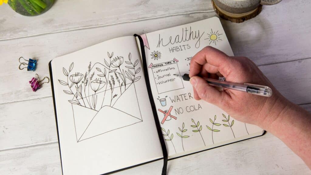 Bullet journal open on self care healthy habits layout pages with hand holding pen. Over shoulder view, fresh white table background, flowers, positive mental health message.