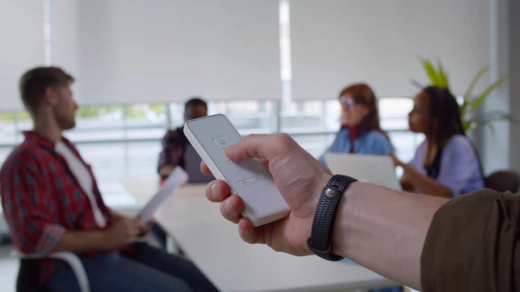 Close up of man hand holds remote control close window blinds in office with multiethnic business team sitting at table and discussing project.