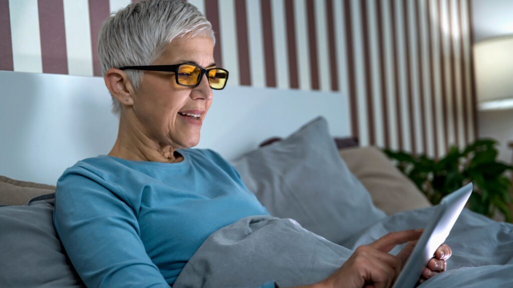 Mature woman wearing blue light blocking glasses with amber lenses, lying in bed before sleep, looking at tablet screen.