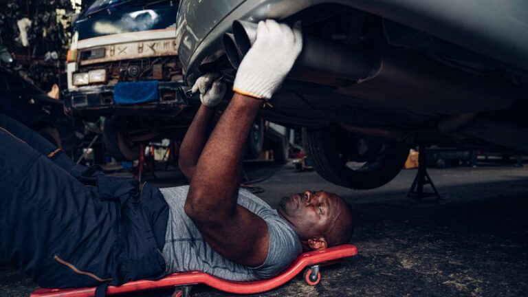 A black male mechanic checking and repairing a vehicle while laying on a wheeled cart underneath.
