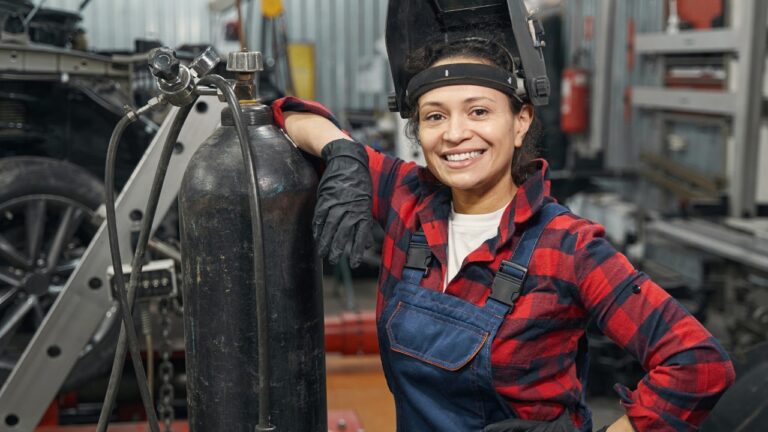 Featured image on 7/12/24. Stock Photo ID: 2164255263. Cheerful woman auto mechanic standing in vehicle repair shop. Female mechanic, auto technician, women in male dominated industry, blue collar woman.
