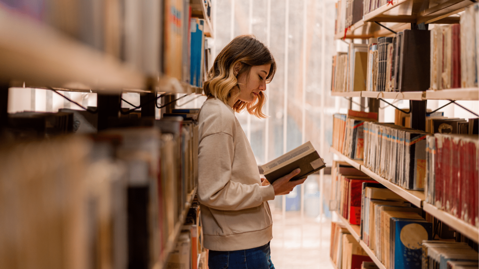 College girl reading and having toughts about the book while standing in the library between the bookshelves.