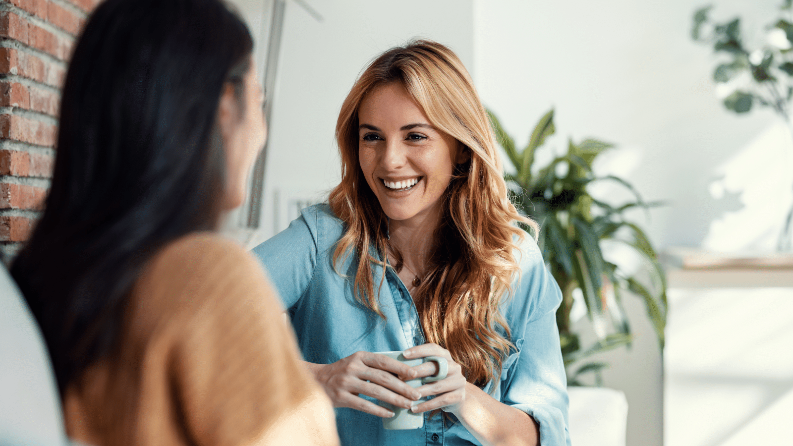 Shot of two smiling young women talking while drinking coffee sitting on couch in the living room at home.