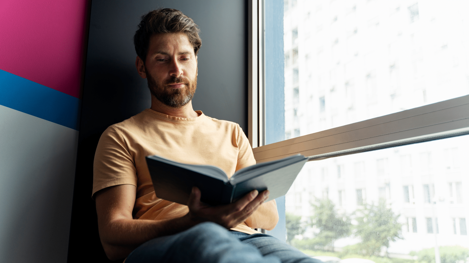 Photo of a man reading book sitting at home near window.