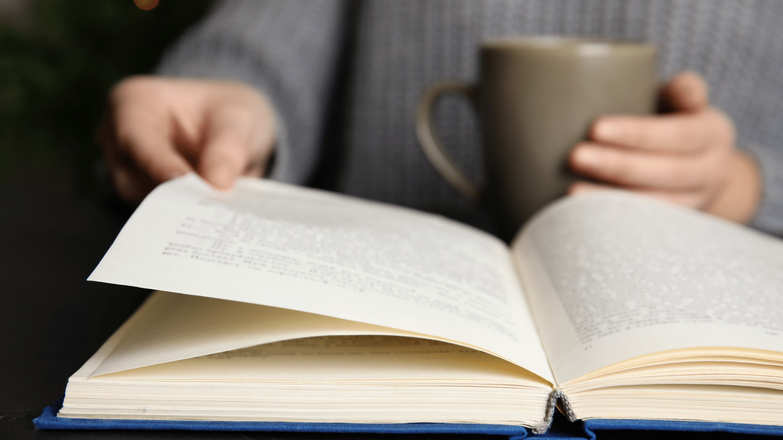 Woman with cup of beverage reading book at table, closeup.