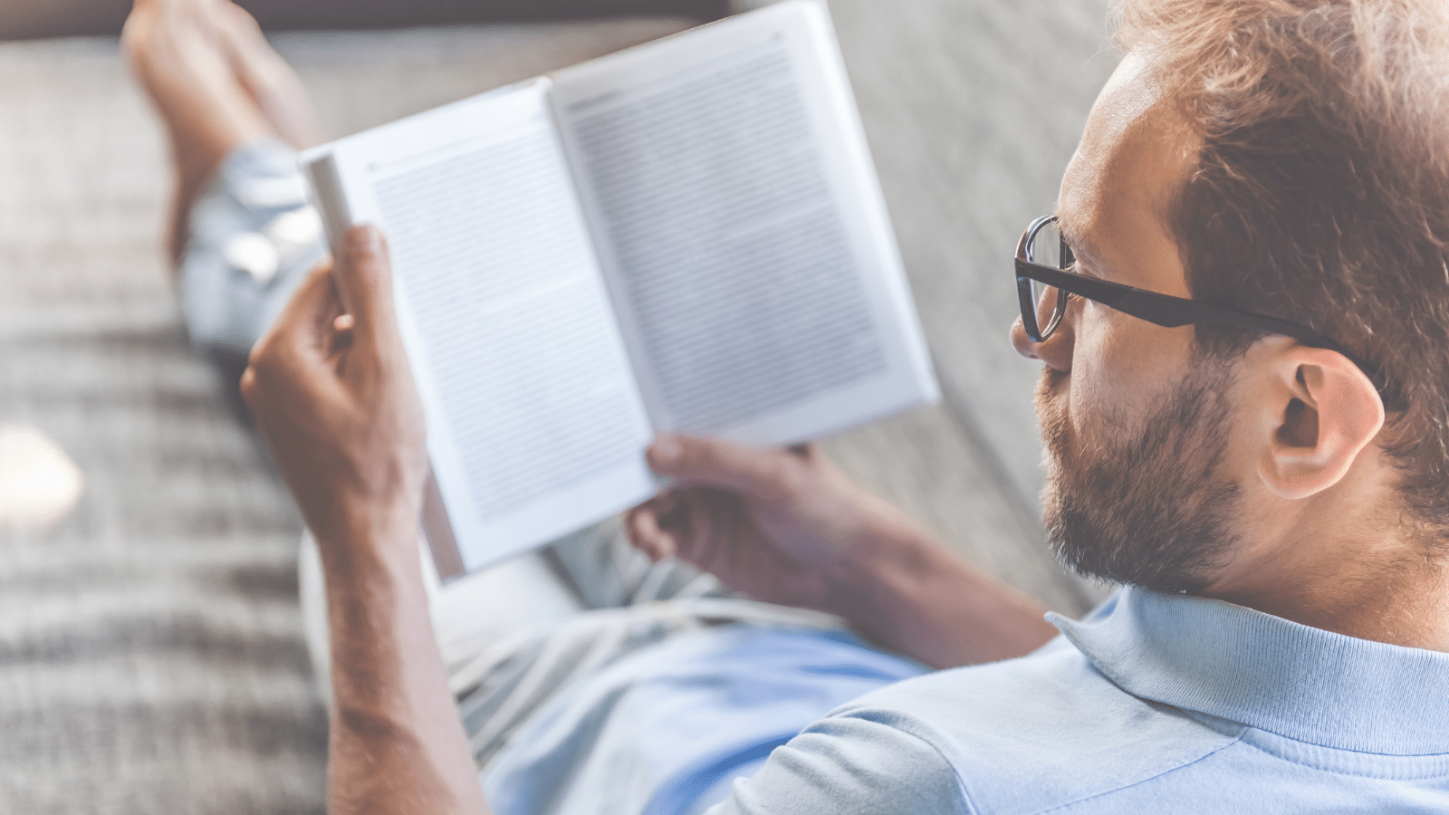 Back view of man in casual clothes and eyeglasses reading a book while lying on couch at home.