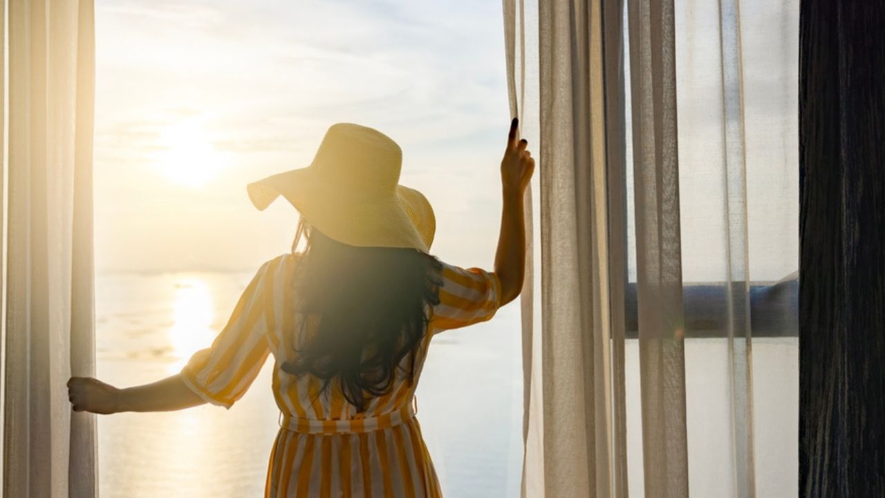 Rear back view woman opening curtains, relaxing and looking beautiful seascape panorama with rays of sun light in holiday on the balcony in high quality hotel feeling happy. Composition of nature