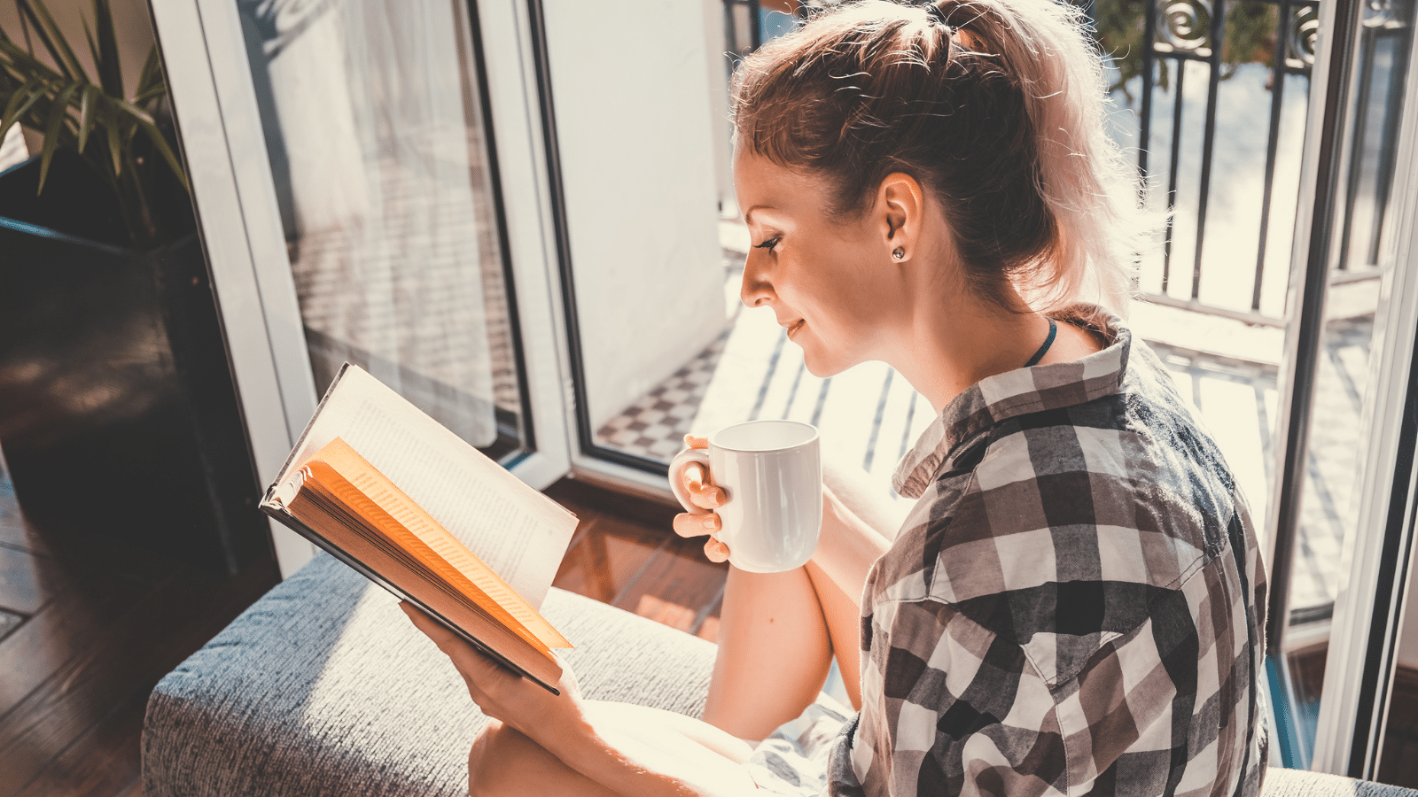 Woman sitting at opened window drinking coffee and reading a book enjoys of rest.
