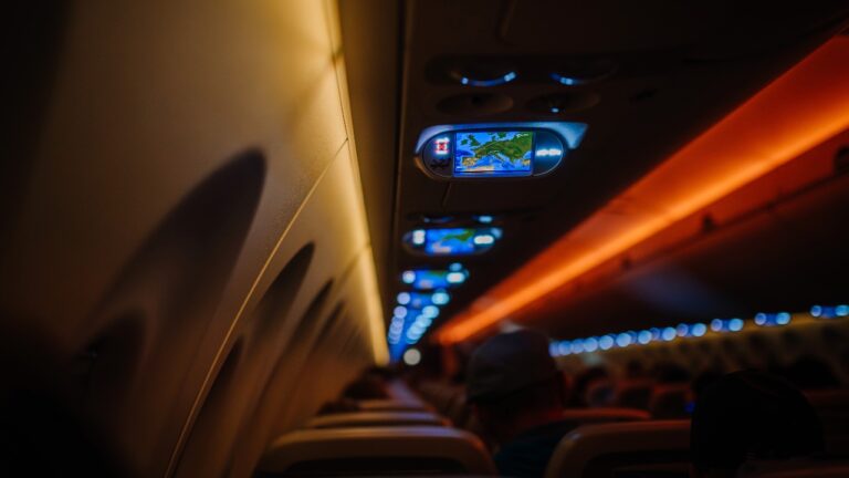 Airplane cabin interior with dim lighting, overhead monitors displaying map, passengers seated, jet lag, dimming, sleeping, 2421760565