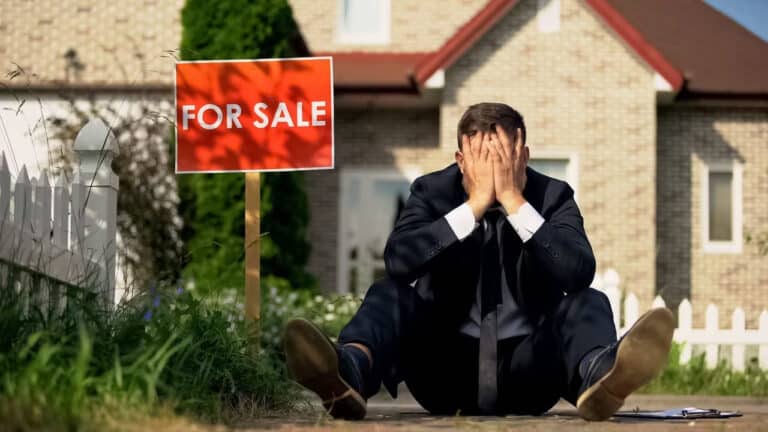 A stressed man sitting besides a for sale sign in a yard.