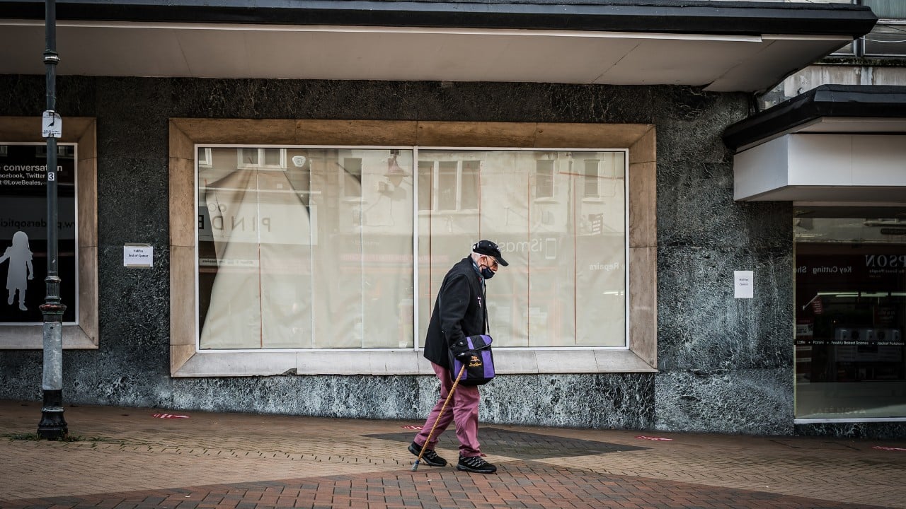 An elderly man walks in front of a closed/shuttered business with a cane. There are blockers in the windows so that nobody can look inside.
