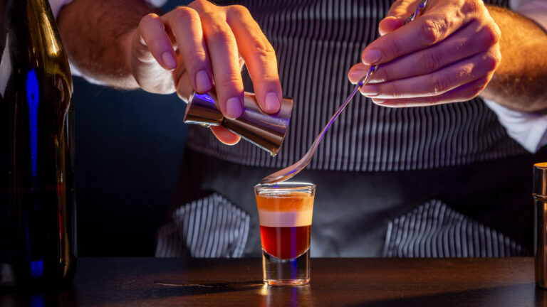 Bartender making B-52 layered cocktail, pouring irish cream from a measuring cup into a shot glass using cocktail spoon