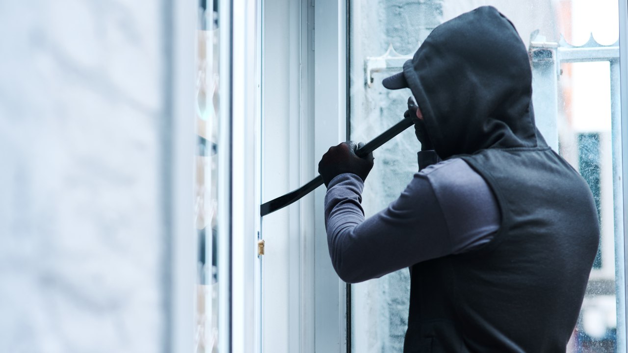 A man dressed in all black, with a hood and face covering, breaking into a home with a crowbar.