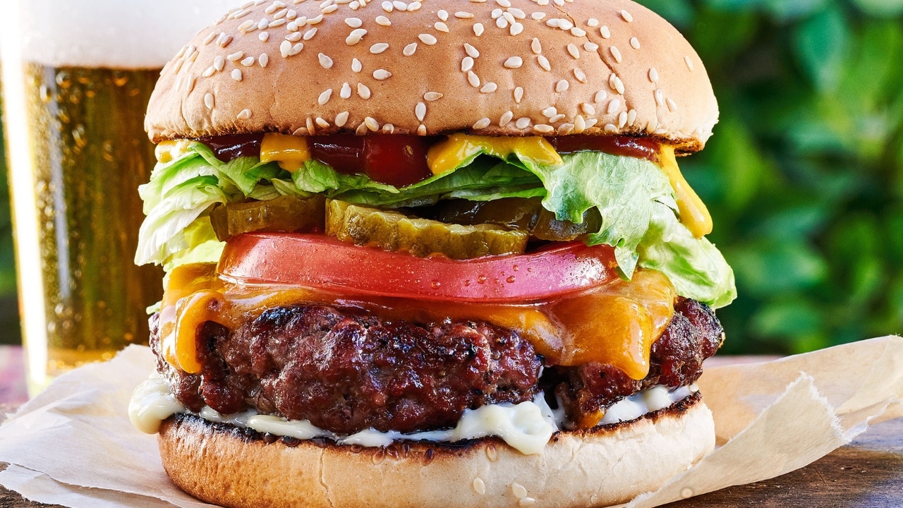 Close view of a loaded cheeseburger with condiments, lettuce, onion, tomato, pickles and cheese on a sesame seed bun.