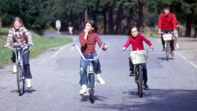 Four people, three girls and one man, riding bicycles, Seattle, Washington, U.S., circa 1980s. Looks like this is probably in a park. Hard to say whether this is a bicycle path or a road that was temporarily closed to cars, but likely one of the two. There appears to be one more cyclist, largely obscured, behind the man.
