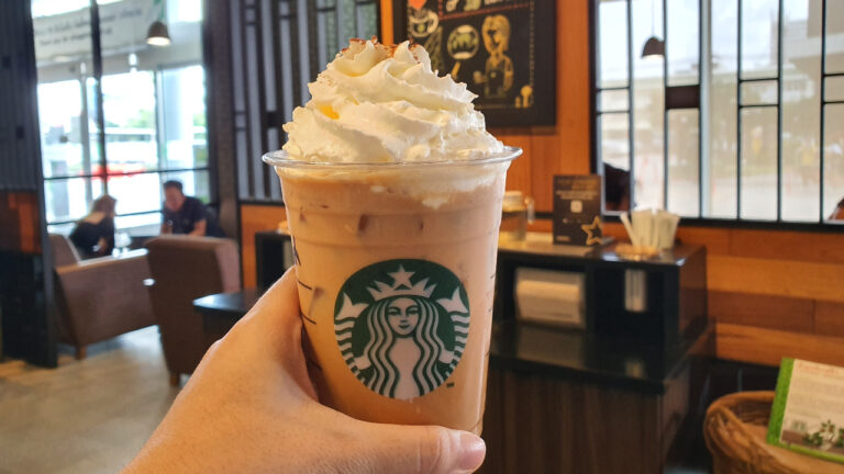 A hand holding a cold pumpkin spice latte in a plastic Starbucks cup with ample whipped cream