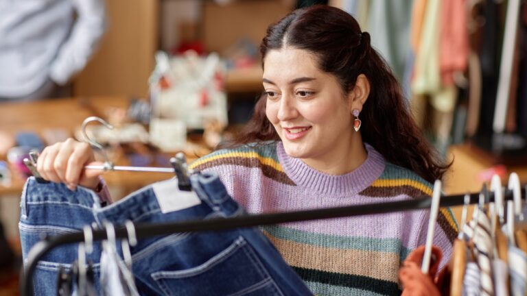 Young woman holding denim jeans on a hanger in a thrift shop. A variety of clothing is in the background on display.