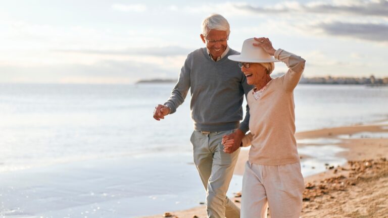 Elderly intelligent couple in love spending time romantically on the beach near the sea, walking holding hands and gently hugging.