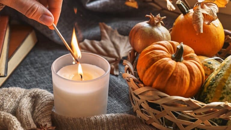 A hand reaching to light a small fogged-glass candle with a match, while comfortable fabric and wicker surrounds the candle on the tabletop. The wicker basket is full of small gourds and pumpkins, leaves and other seasonal items. There are books nearby to show how cozy the area is.