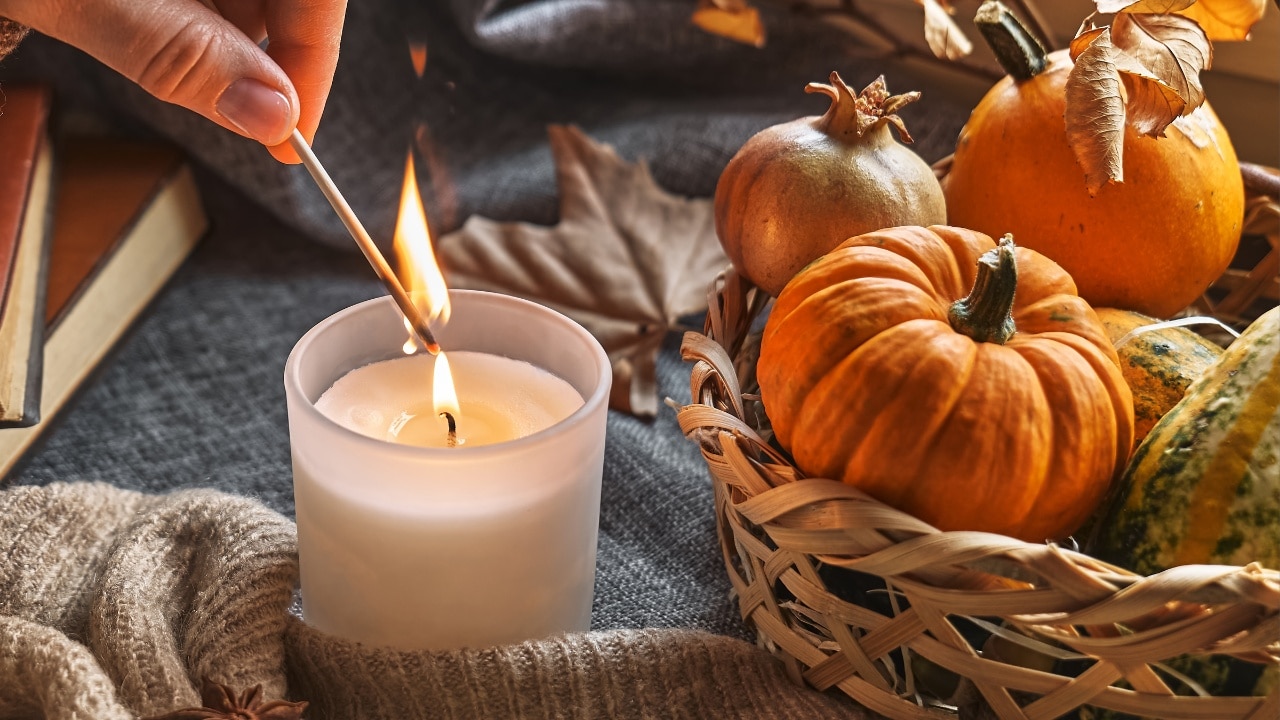 A hand reaching to light a small fogged-glass candle with a match, while comfortable fabric and wicker surrounds the candle on the tabletop. The wicker basket is full of small gourds and pumpkins, leaves and other seasonal items. There are books nearby to show how cozy the area is.