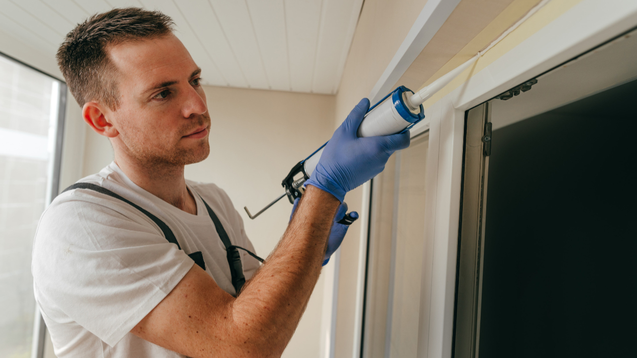 Young man wearing overalls sealing cracks between window and trim using waterproof silicone caulk on the balcony.
