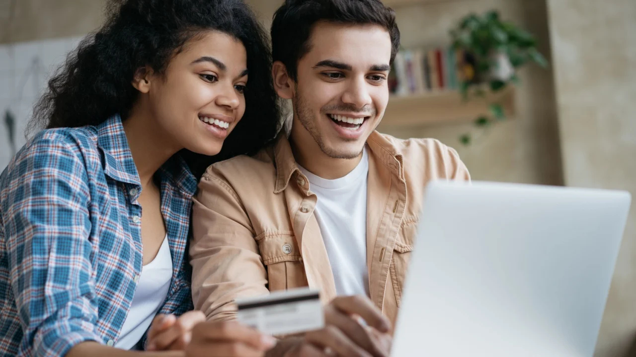 Portrait of beautiful multiracial couple shopping online, using laptop computer. Happy friends ordering food on website. Smiling African America woman holding credit card, looking at digital screen