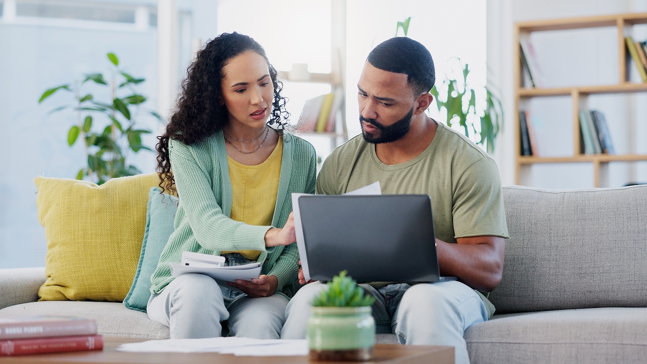 Frustrated couple, finance and documents with laptop on sofa for expenses, bills or financial problems at home. Young, man and woman with paperwork on computer for debt, mortgage or invoices at house