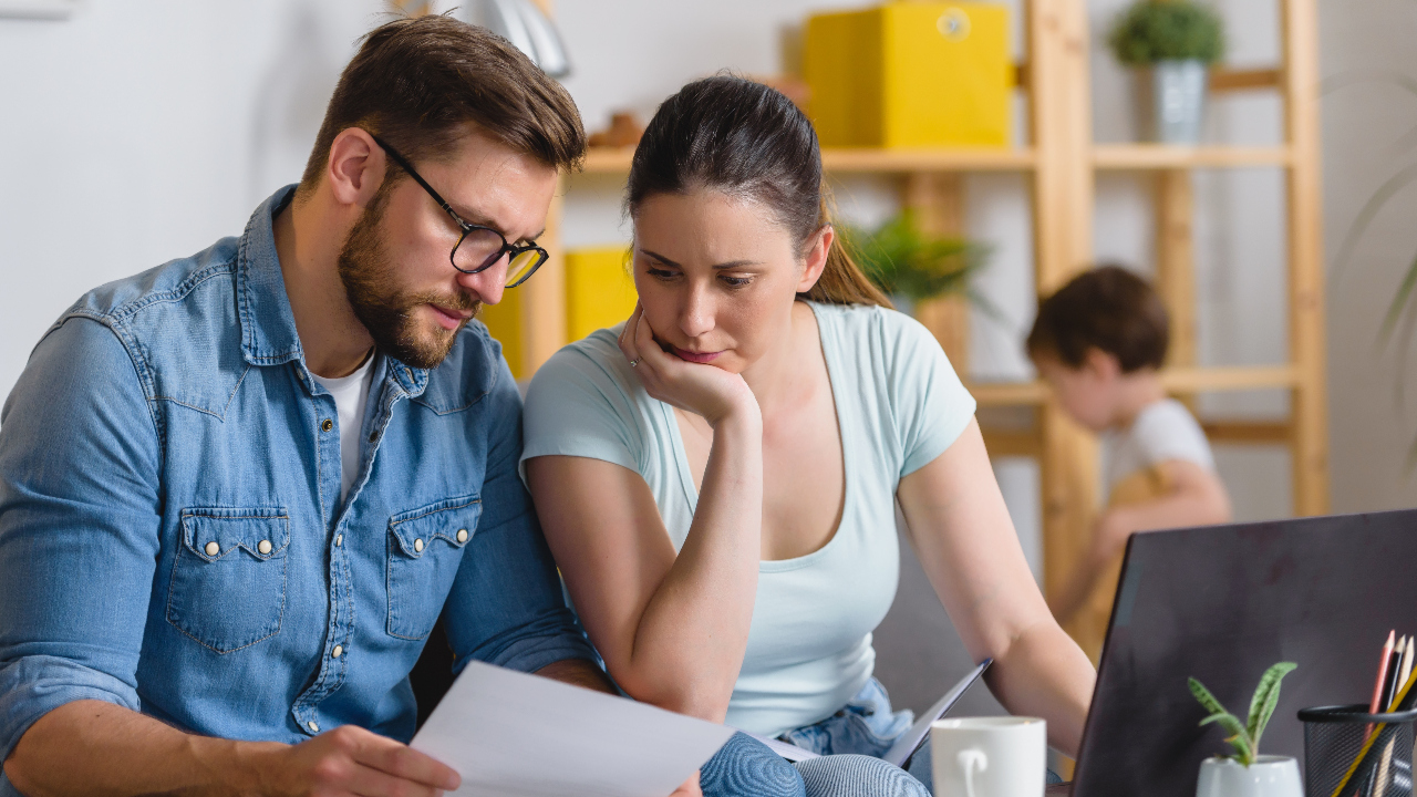 A couple looks worriedly over paperwork while a child plays in the background